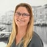 Lächelnde blonde Frau mit lila Brille und grauer Arbeitskleidung vor Schwarz-Weiß-Hintergrund mit Kanal und Gondeln.