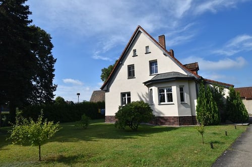 Weißes Haus mit rotem Ziegeldach und Erker, umgeben von grünem Rasen und Bäumen unter blauem Himmel.