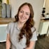 Lächelnde junge Frau mit langen braunen Haaren und grauem T-Shirt mit der Aufschrift "Cut for Friseur".
