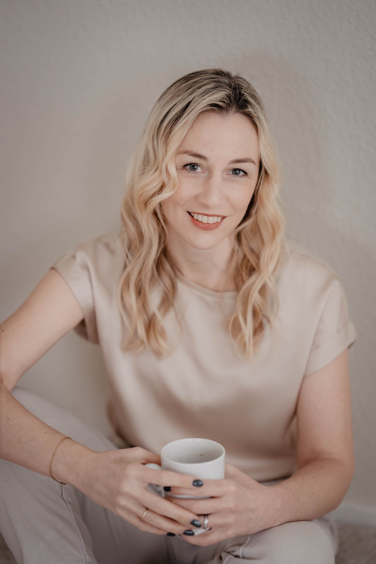 Lächelnde Frau mit blonden, gewellten Haaren hält eine weiße Tasse in beiden Händen und trägt ein beiges Seidenoberteil.