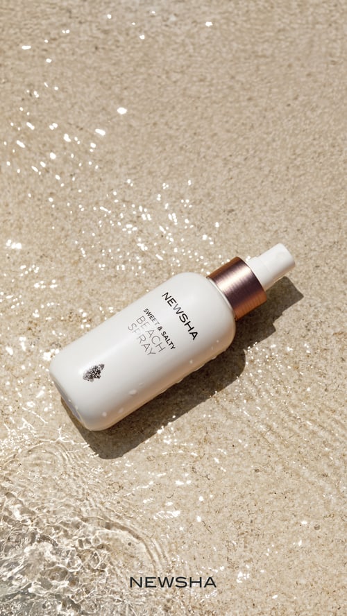 Newsha Sweet & Salty Beach Spray Flasche liegt im flachen, klaren Wasser auf hellem Sand.
