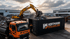 Gelber Bagger belädt einen Hoffmann Metallhandel Container mit Schrott neben einem orangefarbenen LKW auf dem Schrottplatz.