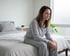 A woman in striped pajamas sits on the edge of a bed with a somber expression.