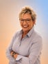 Lachende Frau mit kurzen blonden Haaren und Brille, die eine hellblaue Bluse und eine Smartwatch trägt.
