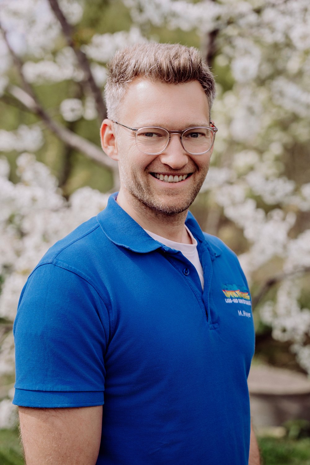 Lächelnder Mann mit Brille und blauem Poloshirt vor einem Baum mit weißen Blüten.