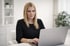 Blonde Frau in schwarzem Kleid tippt auf einem Laptop an einem weißen Schreibtisch.