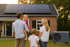 Eine Familie steht vor einem Haus mit Solarmodulen auf dem Dach; das Mädchen hält ein kleines Hausmodell.