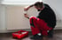 Ein Handwerker in roter Arbeitshose montiert einen digitalen Heizkostenverteiler an einem weißen Flachheizkörper.