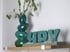 Grüne Vase mit Zweigen und tealfarbene "HPY"-Buchstaben auf einem Holzregal.