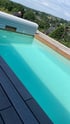 Ein türkisfarbener Außenpool mit Holzterrasse, Poolabdeckung und Blick auf eine grüne Hügellandschaft mit Häusern.