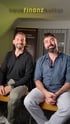 Zwei Männer sitzen auf Stühlen, lächeln in die Kamera, mit dem Text „neuefinanzkultur“ im Hintergrund.
