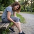 A woman in athletic wear sits on a park bench and grips her lower leg in pain while grimacing.