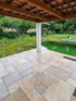 Überdachte Terrasse mit hellen Natursteinplatten neben einem Gartenteich und grüner Bepflanzung.