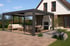 Moderne Terrassenüberdachung mit Glaswänden, Loungemöbeln und Essbereich, angrenzend an ein weißes Haus mit Garten.