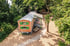 Ein hölzerner Bauwagen mit grünen Fensterläden steht auf einem Waldgrundstück mit Rindenmulch und Bäumen.