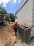 Bauarbeiter und Bagger errichten eine graue Sichtschutzmauer aus Beton an einer hellen Ziegelwand auf einer Erdbaustelle.