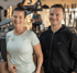 Zwei lächelnde Personen, eine Frau und ein Mann, stehen in einem Fitnessstudio mit Trainingsgeräten im Hintergrund.