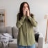 Woman with long brown hair in olive shirt blowing nose with tissue, showing allergy symptoms