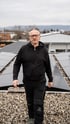 Mann mit grauem Haar, Brille und schwarzem Hoodie geht auf einem Kiesdach mit Solarmodulen.