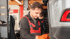 Mechaniker in rotem Overall und orangefarbenen Handschuhen arbeitet an einem Auto in einer Werkstatt.
