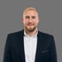 ebastian Hohmann, seriöses Business-Porträt eines Mannes mit blondem Bart im schwarzen Anzug mit weißem Hemd vor grauem Hintergrund.