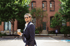 James Lanigan in a suit and sunglasses holds a drink while walking past a brick building and trees.