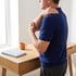 A man in a blue shirt stands by a desk holding his shoulder and elbow, indicating joint pain or muscle tension.