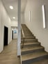 Moderner Flur mit braun gefliestem Boden, einer Treppe mit Metallgeländer und mehreren schwarzen Türen.