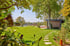 Moderner Garten mit grüner Rasenfläche, Steinpfad, Pool und einem Gartenhaus aus Holz unter schattigen Bäumen.