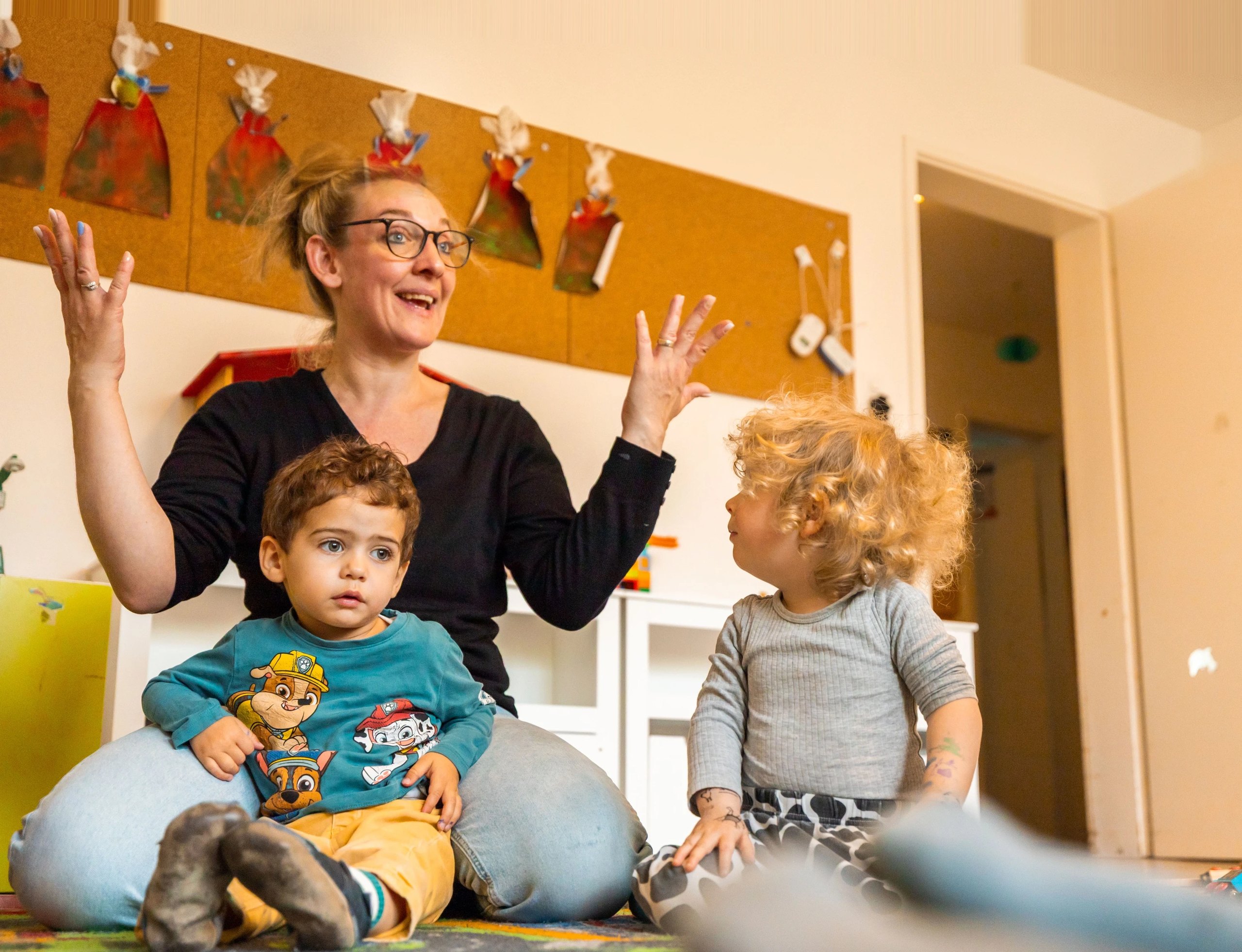 Barbara erzählt im Spielzimmer mit erhobenen Händen, zwei Kleinkinder sitzen vor ihr.