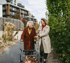 Eine junge Frau begleitet eine ältere Frau mit Rollator auf einem gepflasterten Weg, moderne Gebäude im Hintergrund.