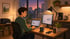 Mann arbeitet abends am Laptop und externen Monitor in beleuchtetem Homeoffice mit Stadtblick.