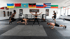 Fünf Personen trainieren in einem Fitnessstudio mit verschiedenen Nationalflaggen an der Wand und einem Boxring im Hintergrund.