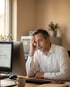 Ein gestresster Mann sitzt vor einem Computer und hält seine Stirn mit der Hand.