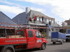 Arbeiter auf dem Dach eines Hauses mit Gerüst; roter Transporter und Anhänger davor geparkt.