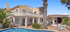 Beigefarbene Villa mit Balkon, Pool, Palmen und einer Terrasse mit Pergola und Sitzgelegenheiten unter blauem Himmel.