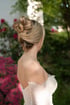 Blonde Braut mit eleganter Hochsteckfrisur und schulterfreiem, weißem Spitzenkleid vor einem Gartenhintergrund.