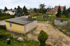 Ein gelbes Gartenhaus mit Flachdach und ein kleines graues Gartenhäuschen auf einem sandigen Grundstück, umgeben von Bäumen und Häusern.