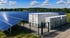 Große Solaranlage auf einer Wiese mit weißen Container-Batteriespeichern und Wechselrichtern hinter einem Zaun.