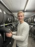 Ein lächelnder junger Mann mit blondem Haar und hellem Pullover steht zwischen Regalreihen voller Autoreifen.