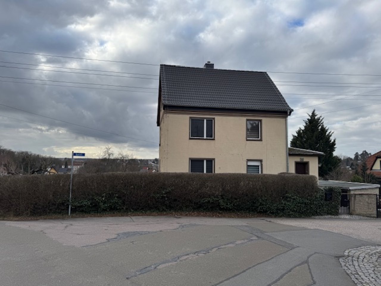 Einfamilienhaus mit Satteldach und heller Fassade hinter einer Hecke an der Straße