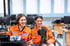 Zwei lächelnde Ingenieurinnen in orangefarbenen Overalls arbeiten an einer schwarzen Roboterhand in einem Computerlabor.