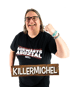 Lachender Mann mit Brille hält ein Holzschild mit der Aufschrift KILLERMICHEL; sein T-Shirt trägt den Text Auswärts Asozial.