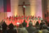 Ein Chor in weißen und türkisfarbenen Gewändern singt vor einem großen Holzkreuz in einer Kirche.