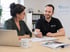 Zwei Personen besprechen Dokumente an einem Schreibtisch mit Laptop, Tasse und "Pflege mit Herz"-Schild.