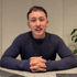 Junger Mann mit dunklem, kurzgeschnittenem Haar, der einen dunkelblauen Rippstrickpullover trägt und lächelt.