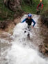 Ein Mann im Neoprenanzug mit Helm rutscht einen Wasserfall hinunter; eine zweite Person steht oben.