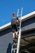 Norman Singer von Sinder & Singer steigt mit Werkzeuggürtel eine Aluminiumleiter an einer grauen Hausfassade hinauf.