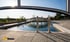 Ein moderner Pool mit Edelstahlleiter, umgeben von Steinfliesen, unter blauem Himmel und mit Bäumen im Hintergrund.