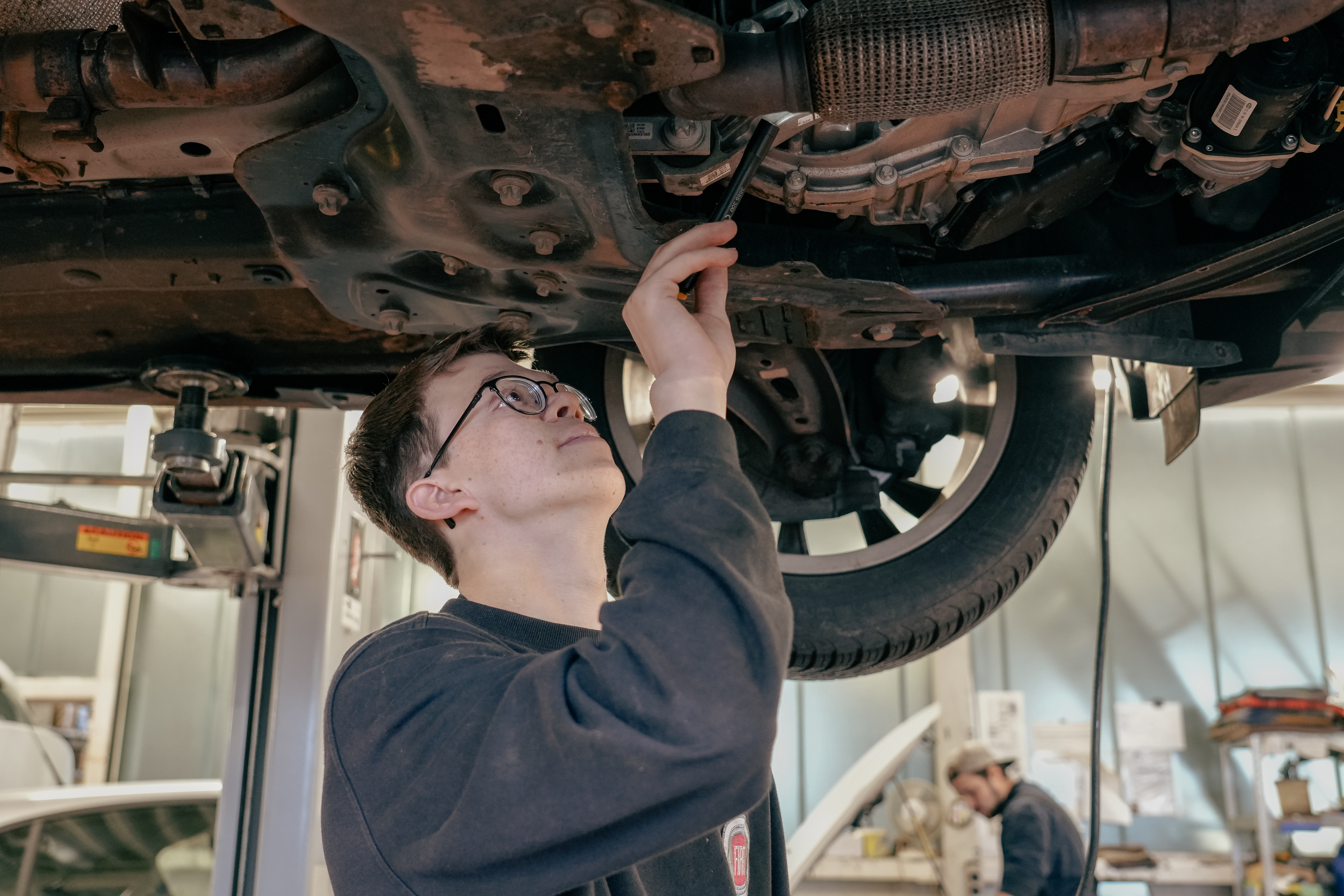 Ein Mann mit Brille inspiziert und repariert die Unterseite eines Fahrzeugs in einer Autowerkstatt.